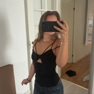 Svart linnetopp med cut out och smala band - Säljer en svart linnetopp med smala axelband och extra banddetaljer över axlarna. Toppen har en snygg cut out framtill och är figurnära i modellen. Perfekt att styla med jeans eller kjol för en trendig look. Helt ny från BikBok! 