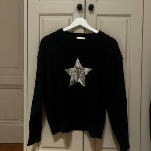 Svart stickad tröja med paljettstjärna - Cool svart stickad tröja från Gina Tricot med en stor silverfärgad stjärna i paljetter på bröstet. Tröjan har rund hals och långa ärmar, perfekt för dig som gillar att sticka ut med lite glitter. Passar dig som vill ha en mysig men ändå trendig look.
