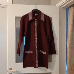 Vinröd långtröja från Giray Triko - Retroinspirerad brun långtröja med rutiga detaljer på krage, knappslå och fickor. Klänningen har stora fickor framtill, långa ärmar och knappar hela vägen. Materialet är stickat och ger en skön känsla. Perfekt för dig som gillar vintagevibbar och unika detaljer. Längd 89cm bredd under armar 56 cm. Storlek M /L