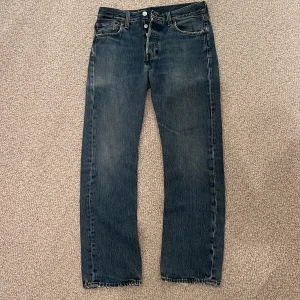W30 L30 Levi's 501 blå jeans straight fit - Klassiska Levi's 501 jeans i blå denim med raka ben och knappgylf. Snyggt slitna detaljer och ikonisk läderpatch bak i midjan. Perfekta för dig som gillar tidlös stil och vill ha ett par jeans som funkar till allt. NYPRIS: Ca. 1400