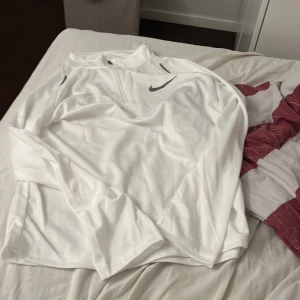 Vit långärmad Nike half zip tröja - Skriv med mig innan ni köper Fräsch vit långärmad tröja från Nike med grått swoosh-tryck på bröstet. Tröjan har en half zip-dragkedja vid halsen och är gjord i mjukt, lätt polyester-material som passar perfekt för träning eller chill. Enkel och stilren design med normal passform. 