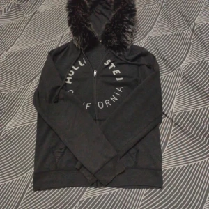 Svart hoodiejacka med fuskpäls från Hollister - Svart hoodiejacka från Hollister med stor fluffig fuskpäls runt huvan och vit text på bröstet. Jackan har dragkedja framtill, snörning vid huvan och två fickor. Perfekt för dig som gillar streetstyle och vill ha något extra mysigt.