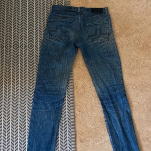 J.Lindberg jeans 32/34 - Säljer ett par blå jeansbyxor från j.lindberg i storlek 32/34. Jeansen har klassisk femficksdesign, raka ben och snygga slitningar. Bakfickorna har dekorativa sömmar och byxorna är tillverkade i denim. Perfekta för en avslappnad och cool stil.