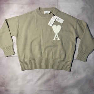 Beige stickad tröja från Ami - Snygg beige stickad tröja från Ami med ribbade muddar och rund hals. På bröstet finns ett vitt hjärta med bokstaven A. Tröjan har en loose passform och är perfekt för dig som gillar stilrena detaljer och mjuka material.