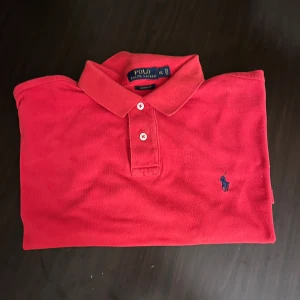 Röd pikétröja Polo Ralph Lauren XL - Snygg röd pikétröja från Polo Ralph Lauren i storlek XL. Klassisk krage, två knappar och den ikoniska blå broderade loggan på bröstet. Tröjan är i bomull och har en normal passform, perfekt för dig som gillar stilren och sportig look.