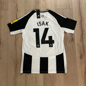 Newcastle United Isak #14 Adidas tröja - Säljer en officiell Newcastle United matchtröja med Isak och nummer 14 på ryggen. Tröjan är svartvit med breda ränder, har korta ärmar och gul sponsorpatch på ena ärmen. Broderat klubbmärke, Adidas-logga och Aeroready-material för skön känsla. Slim fit-modell.