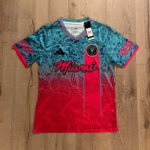 Inter Miami kortärmad fotbollströja - Snygg Inter Miami fotbollströja från Adidas i slim fit. Tröjan har en cool mix av blått och rött med tropiska blad och abstrakta mönster, samt rosa detaljer på ärmar och krage. Materialet är lätt polyester, perfekt för match eller träning. MLS-logga och Miami-tryck på bröstet.