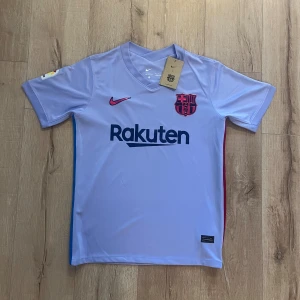 Ljuslila FC Barcelona Nike fotbollströja - Säljer en ljuslila FC Barcelona matchtröja från Nike med korta ärmar och Dri-FIT material. Tröjan har klubbmärke i rött, Rakuten-tryck på bröstet och La Liga-patch på ärmen. Snygga blå och röda detaljer längs sidorna och UNICEF-tryck på ryggen.