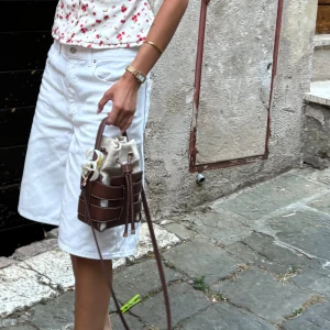 Brun och beige bucket bag med snörning - Säljer en snygg bucket bag i brunt skinn med beige tyginsats och snörning upptill. Väskan har både handtag och lång axelrem, perfekt för att bära crossbody. Stilren design med coola detaljer och kontrastsömmar.