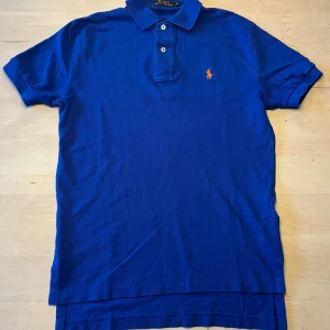 Blå pikétröja från Polo Ralph Lauren XS - Snygg blå pikétröja från Polo Ralph Lauren i storlek XS. Klassisk design med krage, två knappar och orange broderad logga på bröstet. Tillverkad i mjuk bomull som är skön mot huden. Perfekt för dig som gillar stilrena och ikoniska plagg. Väldigt fint skick.