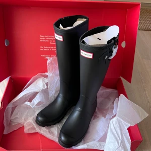 Svarta Hunter stövlar i gummi - Säljer ett par klassiska svarta Hunter stövlar i gummi med rund tå och platt sula. Stövlarna har justerbart spänne vid skaftet och tydlig Hunter-logga framtill. Perfekta för regniga dagar och festivaler. Snygg, stilren design som funkar till många outfits. Oanvända 