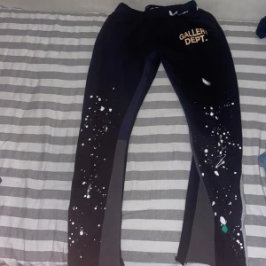 Svarta Gallery Dept. mjukisbyxor - Svarta mjukisbyxor från Gallery Dept. med tryckt logga på höften och unika färgstänk i vitt och grönt längs benen. Byxorna har elastisk midja och inslag av mörkgrå paneler på sidorna. Perfekt för dig som gillar streetwear med konstnärlig vibe.