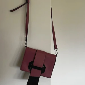 Vinröd axelväska med bred rem - Snygg vinröd axelväska i skinnimitation med bred axelrem och unik dekorativ spänndetalj framtill. Väskan har en praktisk dragkedjeficka på baksidan och stilren, fyrkantig form. Perfekt för dig som vill ha en trendig och funktionell väska.
