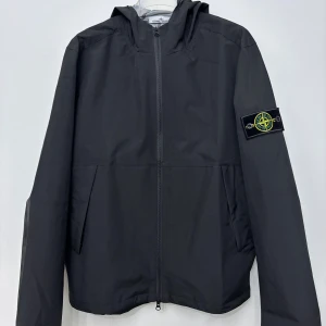 Svart vindjacka från Stone Island - Svart vindjacka från Stone Island med ikonisk patch på vänster ärm. Jackan har huva, hel dragkedja framtill och två sidofickor. Materialet är lätt och syntetiskt, perfekt för blåsiga dagar. Insidan är ljusgrå och jackan har en clean, modern look.