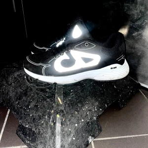 Svarta D sneakers med reflexdetaljer - Säljer ett par svarta D sneakers med feta vita reflexdetaljer och tydlig CD-logga på sidan. Skorna har en chunky vit sula och meshpartier för extra komfort. Snygg design med snörning och sportig vibe, perfekt för dig som gillar statement pieces. Säljer dom för dom ej kommer till andvänding. Inget är sönder på dom. Dom är som helt nya. Pris kan diskuteras.