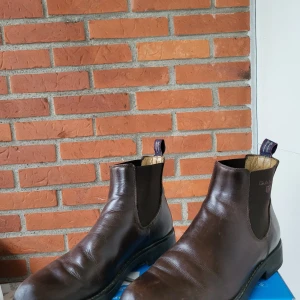 Bruna Chelsea boots från Gant - Snygga Chelsea boots från Gant i mörkbrunt skinn med elastiska sidopaneler och rund tå. Klassisk design med robust svart sula och dragflik baktill med Gant-logga. Perfekta för dig som gillar stilrena och tidlösa skor.storlek 41 men stora i storleken passar 42