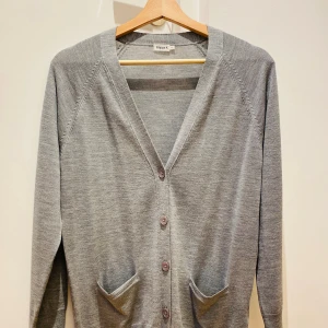 Grå kofta i ull från Filippa K XS - Stilren grå kofta från Filippa K i mjuk ull. Klassisk v-ringning, knäppning framtill med fyra knappar och två fickor. Långärmad modell med ribbade muddar och enkel, clean design som passar perfekt till lager-på-lager.