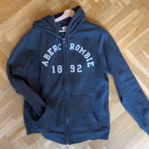 Mörkgrå hoodie från Abercrombie - Mörkgrå hoodie från Abercrombie med vit text och siffror framtill. Tröjan har dragkedja, huva med snörning och två stora fickor. Perfekt för chill dagar och enkel att matcha med jeans eller joggers.