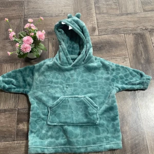 Turkos fleece hoodie med djurdetaljer - Supermjuk turkos hoodie i fleece med djurinspirerade detaljer på huvan, som öron och tänder. Tröjan har ett diskret mönster, stor känguruficka framtill och långa ärmar. Perfekt för dig som gillar mysiga och lekfulla plagg.