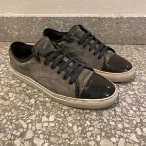 Lanvin - Snygga gråa sneakers från lanvin med ovandel i mocka och blanka detaljer på tå och häl. Skorna har svarta snören och en vit, platt sula som ger en clean look. Perfekta för dig som gillar en stilren och urban vibe. Skick 8/10. Skriv vid frågor eller prisförslag!😁