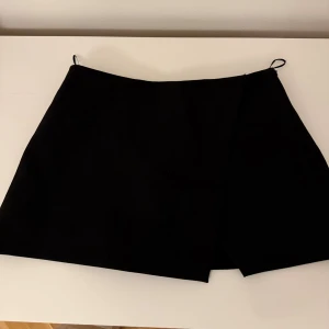 Svart kjol från Zara - Stilren svart omlottkjol från Zara i kort modell. Kjolen har shorts inbyggt inuti vilket är super skönt/smidigt. Aldrig använd och prislappen kvar!