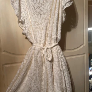Vit spetsklänning med knytband - Säljer en vit klänning med vacker spets och knytband i midjan. Klänningen har korta ärmar och ett fint fall, perfekt för dig som gillar en feminin och bohemisk stil. Spetsdetaljerna ger en extra touch av elegans. Användes endast på studenten. Köptes för 500kr