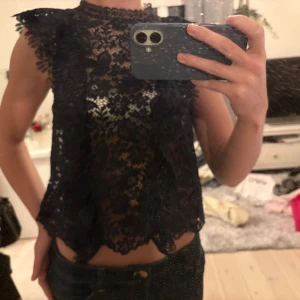Topp Zara - Säljer en svart blus i spets med blommigt mönster och volangdetaljer vid axlarna. Blusen är ärmlös, har hög hals och är croppad med snygg scallop-kant nedtill. Från Zara!