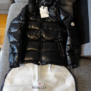 Svart Moncler pufferjacka med huva - Snygg svart pufferjacka från Moncler med glansig finish och ikonisk logga på ärmen. Jackan har huva, dragkedja framtill och praktiska fickor med dragkedja. Insidan har en cool serietidningsetikett. Perfekt för dig som vill ha en stilren och varm jacka. Ingår också dustbagen för jackan