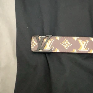 L och V monogram skärp i skinn - Säljer ett brunt L och V bälte med klassiskt monogrammönster och L och V-logga på spännet. Bältet är tillverkat i äkta skinn och har detaljer i beige och guld. Perfekt accessoar för dig som vill ha en lyxig och ikonisk look.