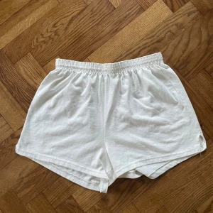 Vita Soft Shorts med resårmidja - Säljer ett par klassiska vita Soft Shorts från Soffe med bred resår i midjan och kort, loose passform. Tillverkade i mjuk bomull, perfekta för chill dagar eller träning. Diskret slits i sidorna för extra rörelsefrihet. Enkla och stilrena, passar dig som gillar basic.