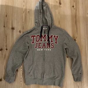 Grå hoodie från Tommy Jeans med dragkedja - Snygg grå hoodie från Tommy Jeans med stort rött och vitt tryck på bröstet. Tröjan har huva med svarta snören, dragkedja framtill och två fickor. Perfekt för chill dagar och streetstyle.