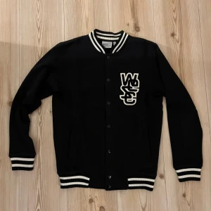 Collegejacka från WESC - strl.XS - Svart collegejacka från WESC i storlek XS med vita ränder på krage, ärmslut och nederkant. Jackan har tryckknappar framtill och en stor vit WESC-logga på bröstet. Snygg och sportig stil, perfekt för dig som gillar streetwear.