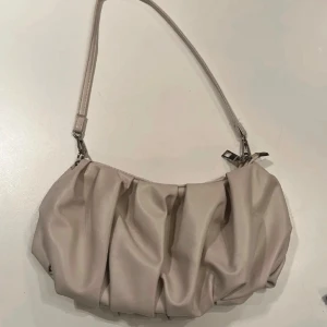 Beige axelväska med veckad design - Snygg beige axelväska med veckad och rynkad design. Väskan har ett smalt axelband och stängs med dragkedja. Materialet är mjukt och har en lyxig känsla, perfekt för att lyfta din outfit. Insidan är mörk och har ett rymligt fack för dina essentials.