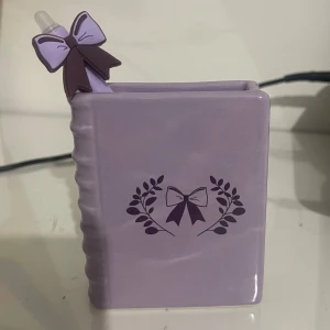 Bokformd behållare  - En lila bokformad behållare med en dekorativ rosett och bladmotiv. Perfekt som pennställ eller dekoration för skrivbordet. Söt och stilren design som passar ungdomar och unga vuxna som gillar kawaii eller pastellstil.