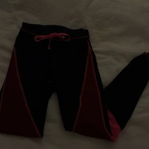 Svarta och rosa träningsleggings - Snygga svarta leggings med rosa detaljer och snörning i midjan. Perfekta för träning eller chill, med en tight passform och stretchigt syntetmaterial som ger bra rörelsefrihet. De har coola paneler i rosa längs benen som ger en sportig vibe.