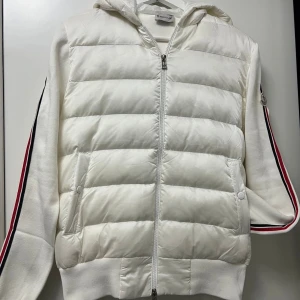 Vit Moncler Cardigan med huva - Säljer en snygg vit Cardigan från Moncler med huva och dragkedja framtill. Jackan har quiltad front, stickade ärmar med röda och svarta ränder samt Moncler-logga på armen. Perfekt för dig som gillar exklusiv streetstyle och vill ha något som sticker ut.