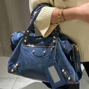 Blå Balenciaga handväska i skinn - Snygg blå handväska från Balenciaga i skinn med guldiga nitar och dragkedjor. Väskan har flätat handtag, spegel och flera fack med dragkedja. Perfekt för dig som vill ha en statementväska med coola detaljer.