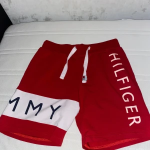 Röda shorts Tommy Hilfiger M - Snygga röda shorts från Tommy Hilfiger i storlek M. De har vit snörning i midjan, stora tryck med loggan på benen och en vit panel med text. Perfekta för sommaren och har en sportig vibe. Materialet är mjukt och skönt, troligen bomull.