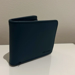 Blå plånbok från Pull&Bear - Snygg blå plånbok från Pull&Bear i stilren design med flera kortfack och en praktisk dragkedja för mynt. Tillverkad i slätt skinnmaterial och har en kompakt, fyrkantig form som är enkel att ta med sig. Perfekt för dig som vill ha koll på kort och cash.