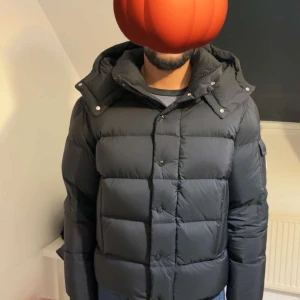 Svart Moncler pufferjacka med huva - Svart pufferjacka från Moncler med avtagbar huva, stora quiltade sektioner och ikonisk logga på ärmen. Jackan har både dragkedja och knappar framtill, samt snygga detaljer i rött och blått på insidan. Perfekt för kalla dagar och riktigt stilren.