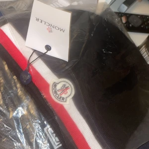 Svart Moncler mössa med röd-vit rand - Svart mössa från Moncler med klassisk röd och vit rand längs kanten och broderad Moncler-logga framtill. Stickad i mjukt material som håller dig varm och snygg. Perfekt accessoar för att lyfta din vinterstil.