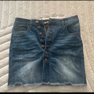 Blå jeanskjol med knappar - Snygg blå jeanskjol från Indie Rose med knappar framtill och fransig nederkant. Klassisk femficksmodell med bälteshällor och slitna detaljer för en cool look. Perfekt till sneakers eller boots.