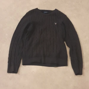 Mörkblå kabelstickad tröja från GANT - Mörkblå kabelstickad tröja från GANT med rund hals och diskret logga på bröstet. Tröjan har långärmad passform och ribbade muddar vid ärmslut och nederkant. Perfekt för dig som gillar klassisk stil med en modern touch.