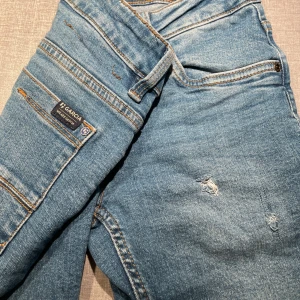 Blå Garcia jeans med slitningar, strl 140 - Säljer ett par ljusblå jeans från Garcia i storlek 140. Jeansen har coola slitningar och detaljer på framsidan, klassisk femficksdesign och kontrastsömmar. Perfekt för dig som gillar en avslappnad och trendig look.
