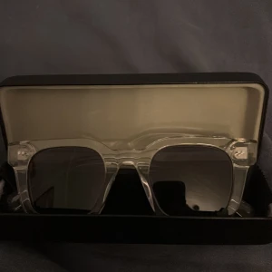 Transparenta solglasögon All Cap One - Säljer ett par stilrena solglasögon från All Cap One med transparent, fyrkantig båge och mörka linser. Snygga detaljer i silver på skalmarna. Kommer med original svart fodral med logga. Perfekt för dig som vill sticka ut med en modern look.