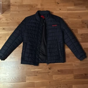 Svart quiltad pufferjacka från Hugo boss - Svart pufferjacka från Hugo boss med röd logotyp på bröstet. Jackan har quiltad design, dragkedja framtill och praktiska fickor. Insidan är slät med en innerficka som har röd dragkedja. Perfekt för kyliga dagar och har en modern, stilren look. Säljer den då den är för stor för mig. Fick den i present förra året aldrig använd. Nypris 3000kr. Pris går att diskutera!