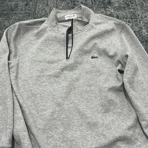 Grå half zip tröja från Lacoste - Snygg grå långärmad tröja från Lacoste med klassisk krokodil-logga på bröstet. Tröjan har half zip-dragkedja och en stilren, enkel design. Regular fit och mjukt material som känns skönt mot huden. Perfekt för en clean och sportig look.