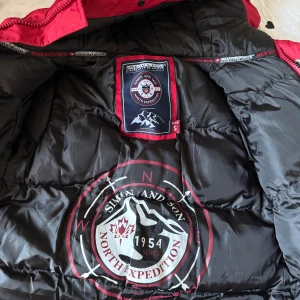 Röd pufferjacka från Simon and Son - Säljer en röd pufferjacka från Simon and Son North Expedition med huva och flera praktiska fickor med dragkedja. Jackan har coola svarta detaljer och tryck på insidan. Perfekt för kalla dagar och har en riktigt schysst streetstil.