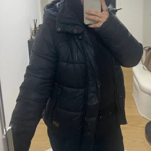 Svart pufferjacka från G-Star RAW Herr - Svart pufferjacka från G-Star RAW med huva, dragkedja och snörning vid kragen. Jackan har quiltad design, ribbade muddar och två fickor med dragkedja. Märkesdetaljer på bröstet och bak. Perfekt för kalla dagar och har en modern, urban vibe.