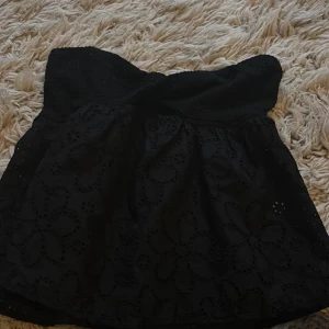 Svart axelbandslös topp med spets - Svart kort axelbandslös klänning med broderade blommor och spetsdetaljer. Överdelen är smockad för en bekväm passform och kjolen har ett fint blommigt mönster i spets. Perfekt för dig som gillar en feminin och trendig stil.